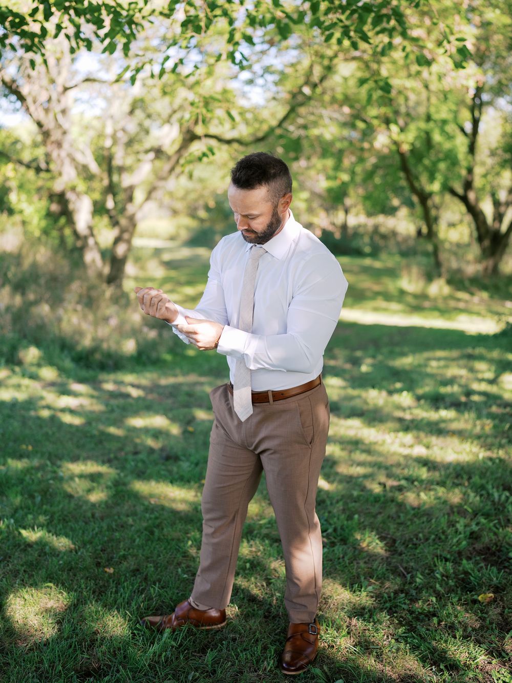 Groom in white shirt and tan pants adjusting cufflinks outdoors in a lush green orchard setting.