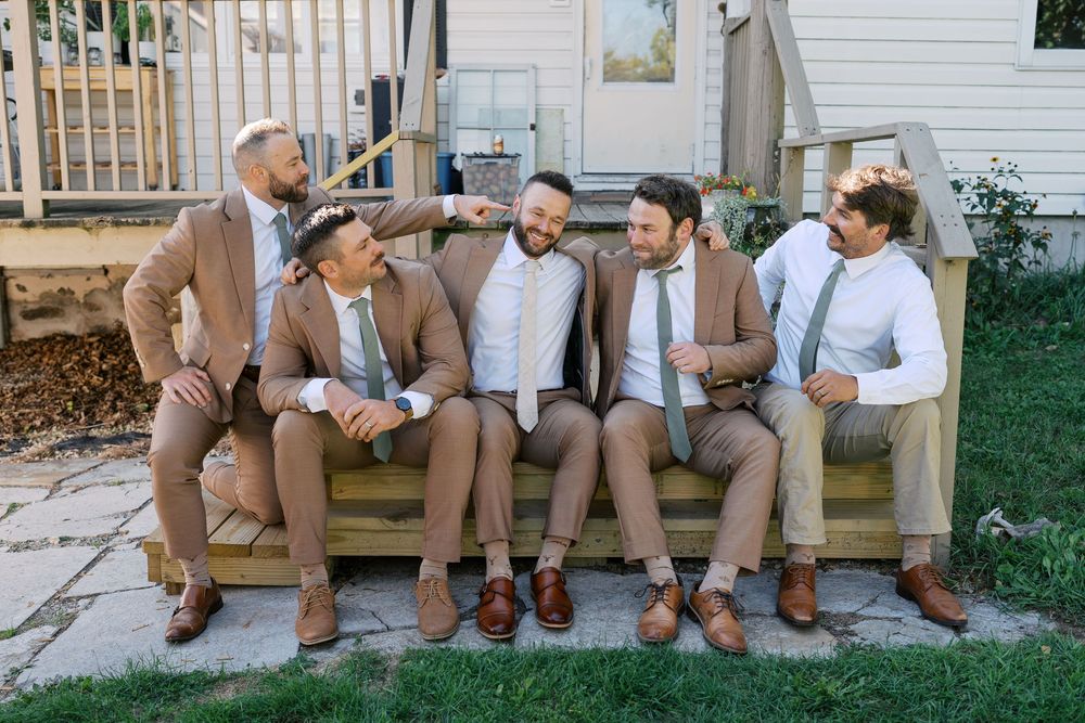 Five groomsmen in tan suits and brown shoes sit on a wooden bench outdoors, laughing together at a wedding.