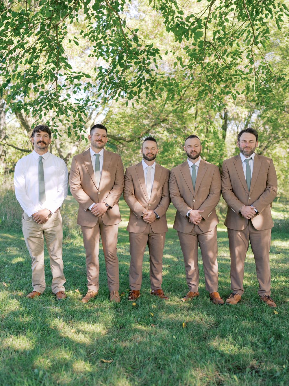 Five groomsmen in matching tan suits and sage ties pose outdoors under green trees on a sunny wedding day.
