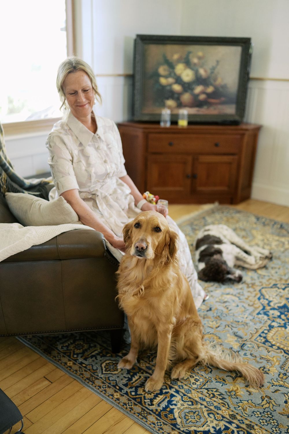 A smiling blonde woman in a white dress sits on a leather sofa petting a golden retriever on a blue patterned rug.