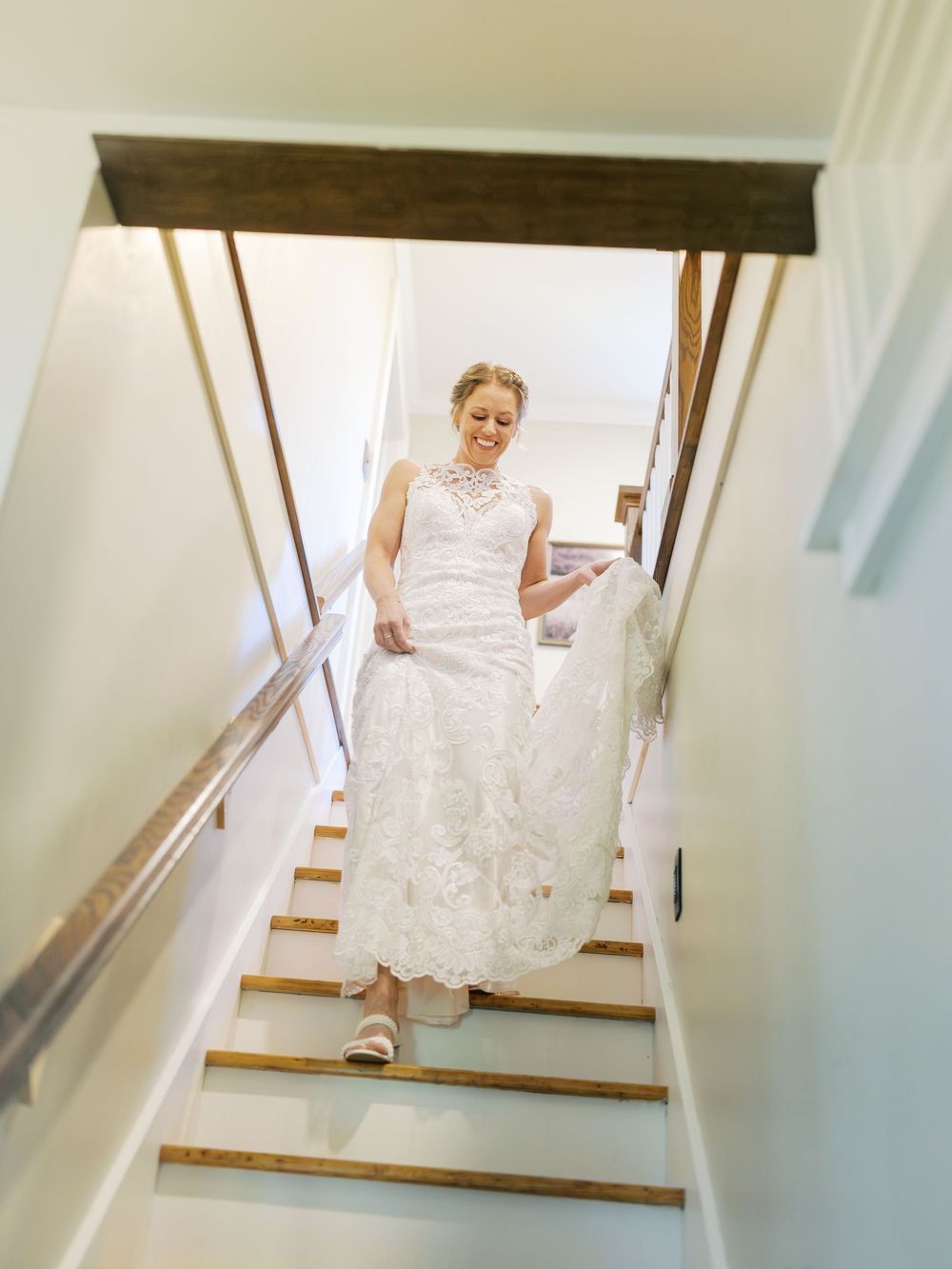 Bride in white lace wedding gown descending wooden staircase, lifting dress hem, smiling softly in bright interior.