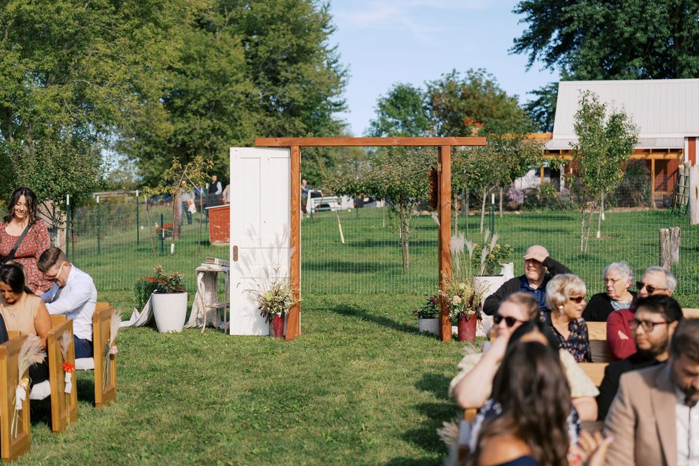 Outdoor wedding ceremony setup with wooden arch, floral decor, and guests seated on benches in a lush green garden.