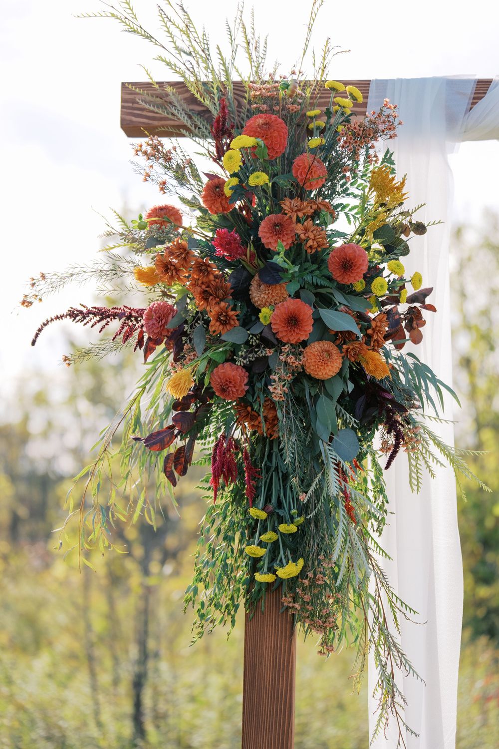 Autumn wedding arch floral arrangement with orange dahlias, greenery, and burgundy accents on a wooden post.