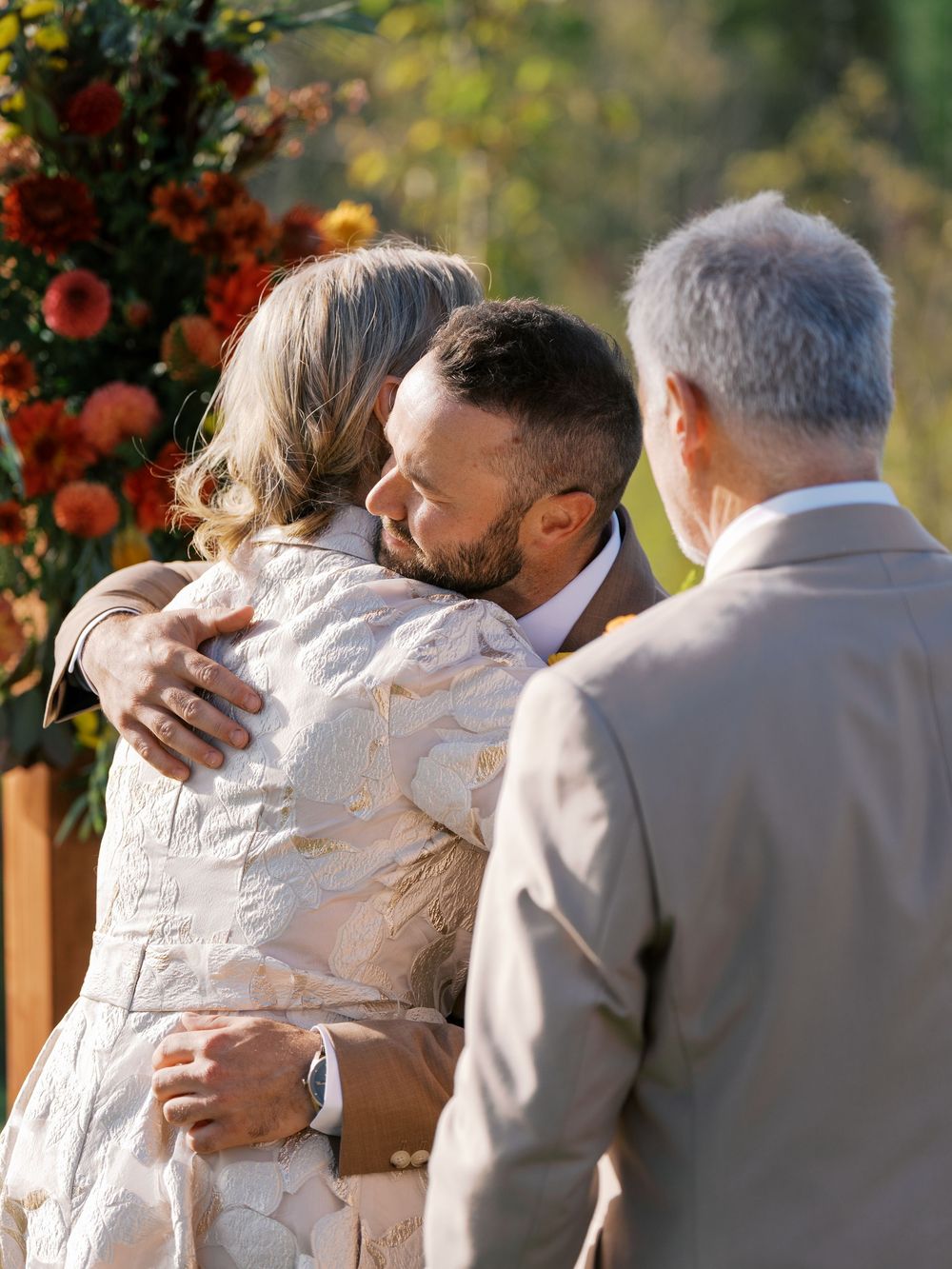 Groom embracing a woman in a floral lace dress at an outdoor fall wedding ceremony with autumn floral arrangements.