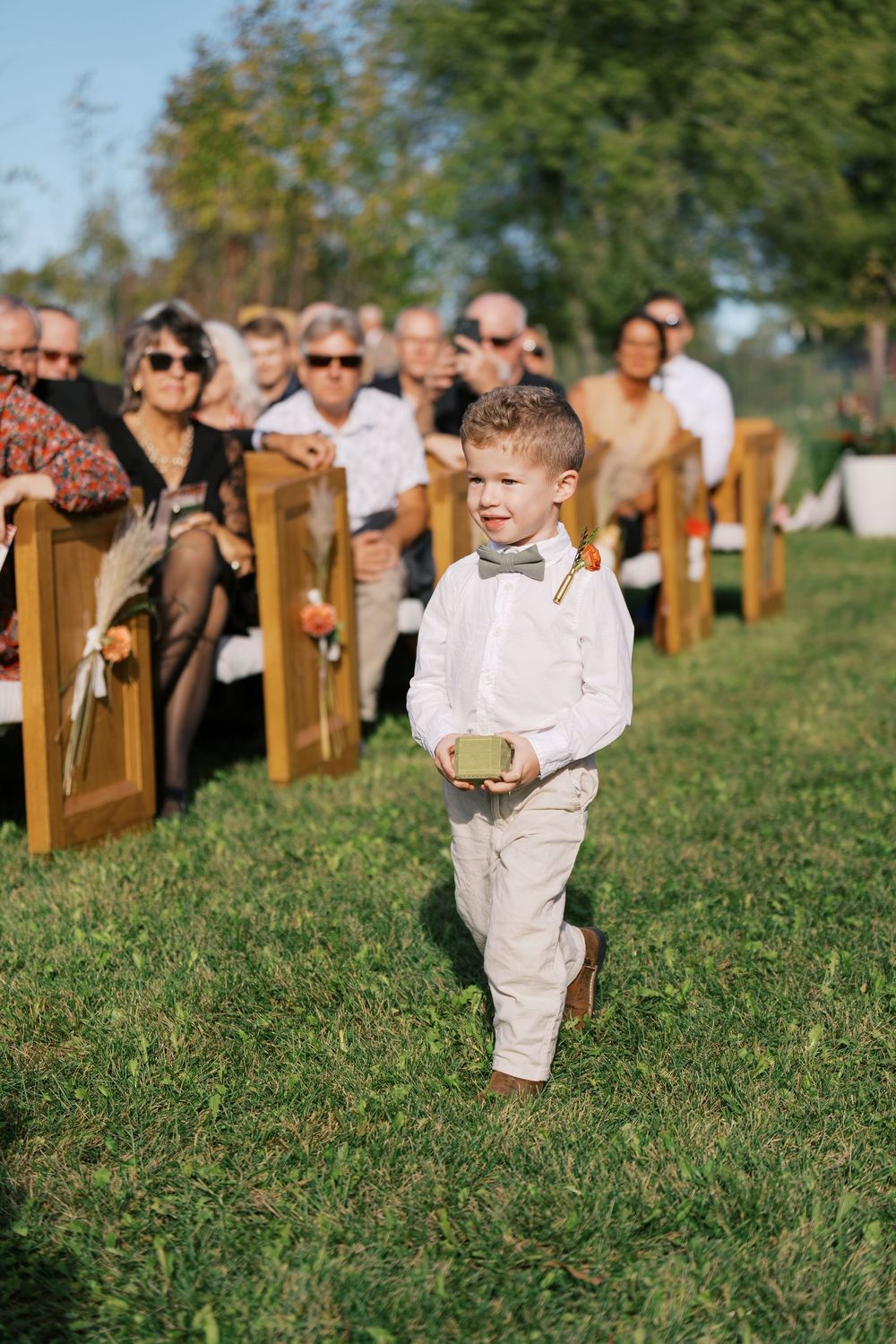 A young ring bearer in white shirt and suspenders walks down the aisle at an outdoor wedding ceremony.