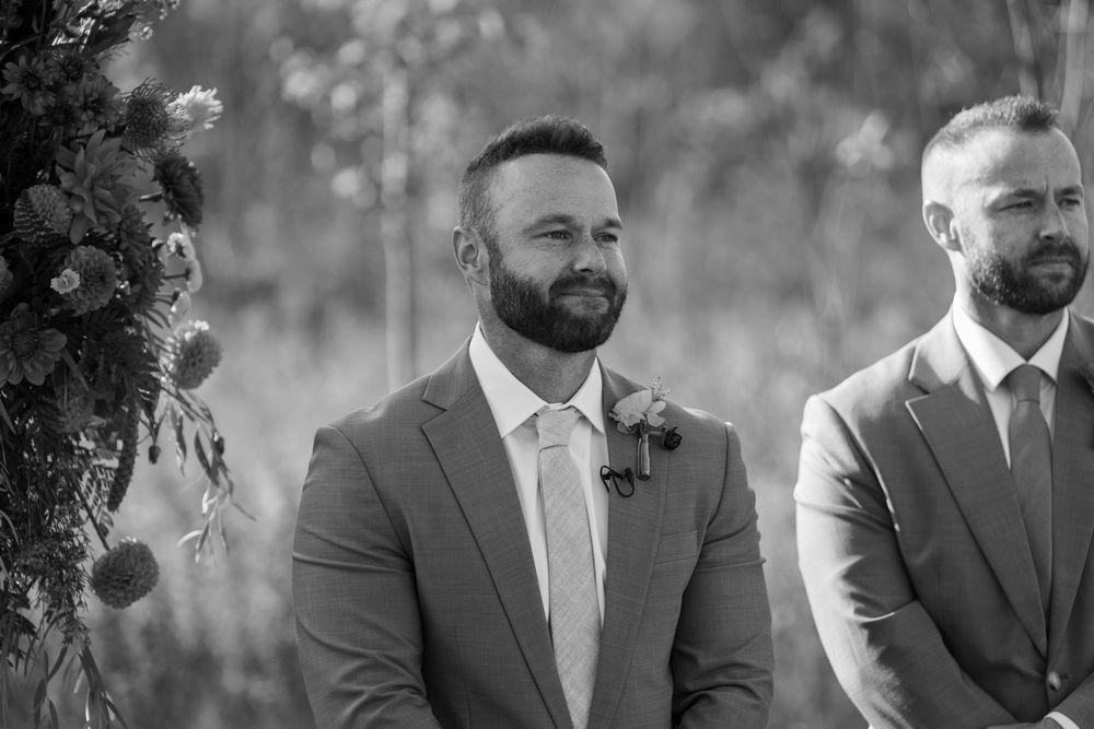 Groom smiling at altar in gray suit with boutonniere during outdoor wedding ceremony, black and white photo.