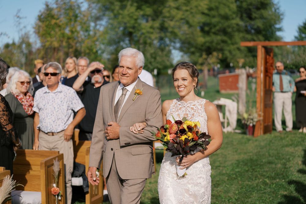Bride walking down outdoor aisle with father, holding fall bouquet, guests seated, wooden arch in background.