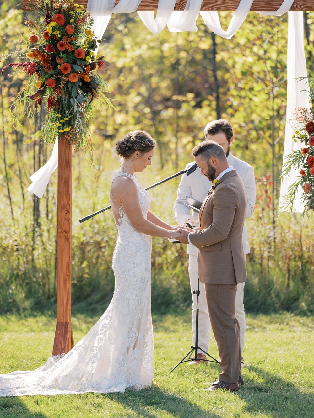 Outdoor fall wedding ceremony with bride and groom exchanging vows under a wooden arch adorned with autumn floral arrangements.