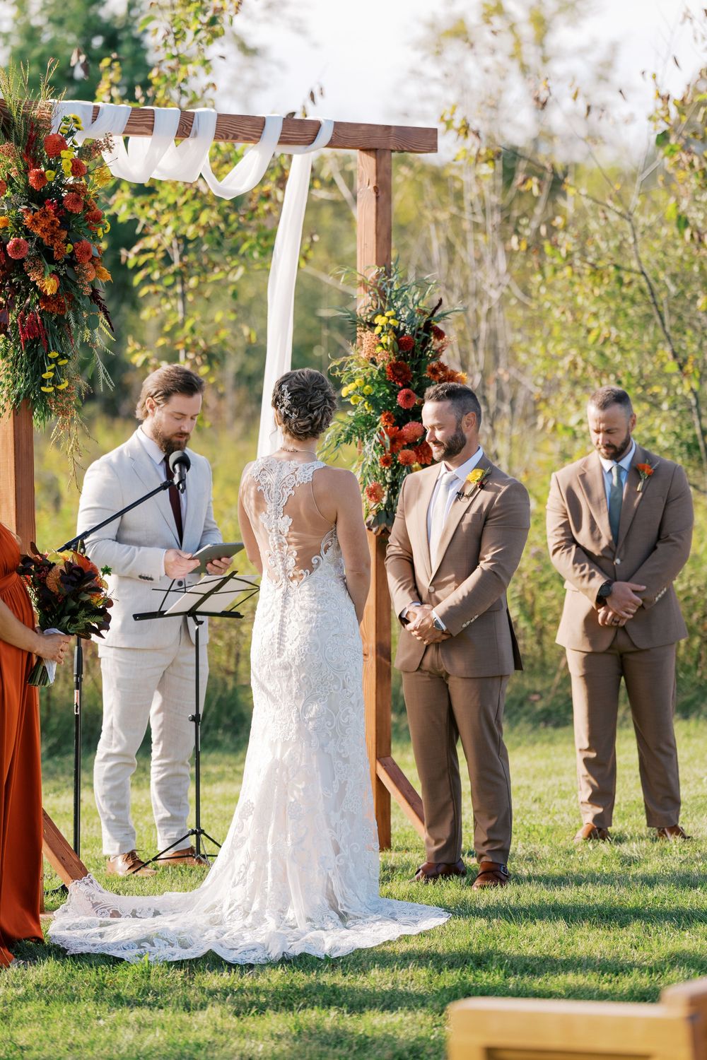 Outdoor fall wedding ceremony with bride and groom at wooden arch decorated with orange and red florals.
