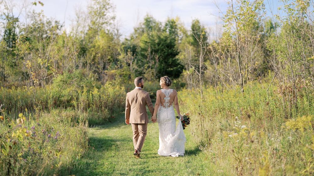 Bride and groom walking hand in hand through a lush green meadow path surrounded by trees on their wedding day.