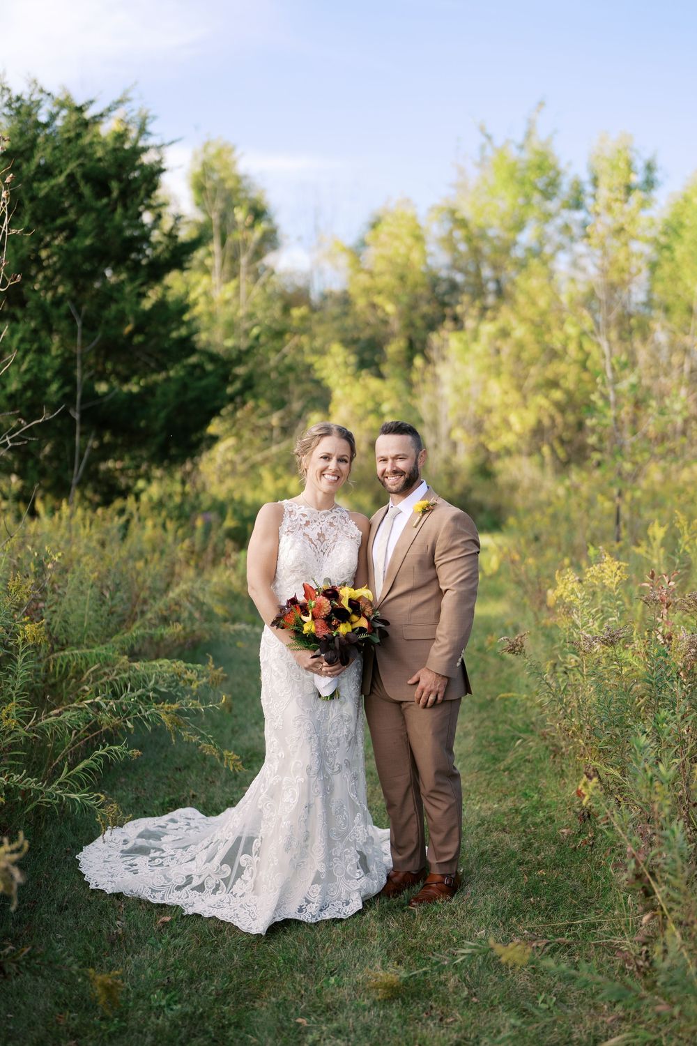 Bride and groom pose together outdoors in lush greenery, she in lace gown with bouquet, he in tan suit.