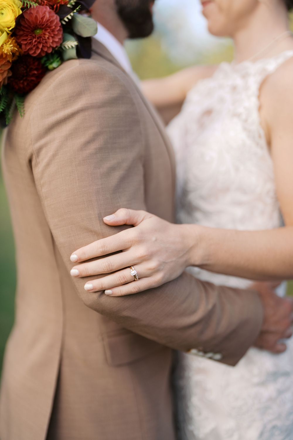 Bride in lace gown rests hand with diamond ring on groom's tan suit jacket, with fall floral bouquet.