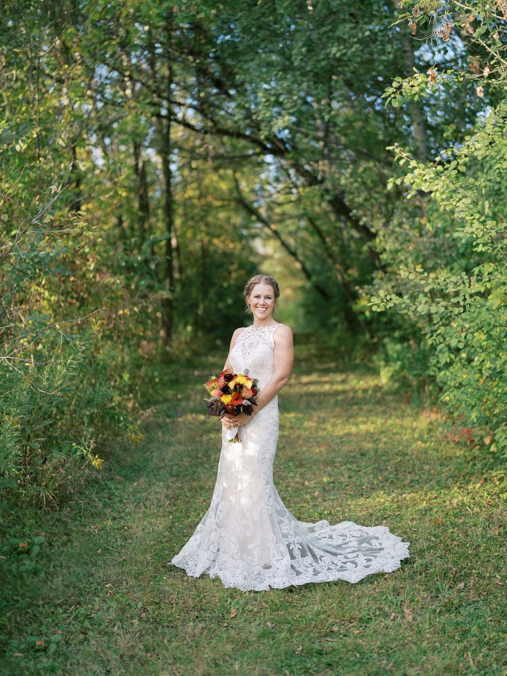 Bride in lace mermaid wedding gown holding fall bouquet of sunflowers stands on grassy path surrounded by green trees.