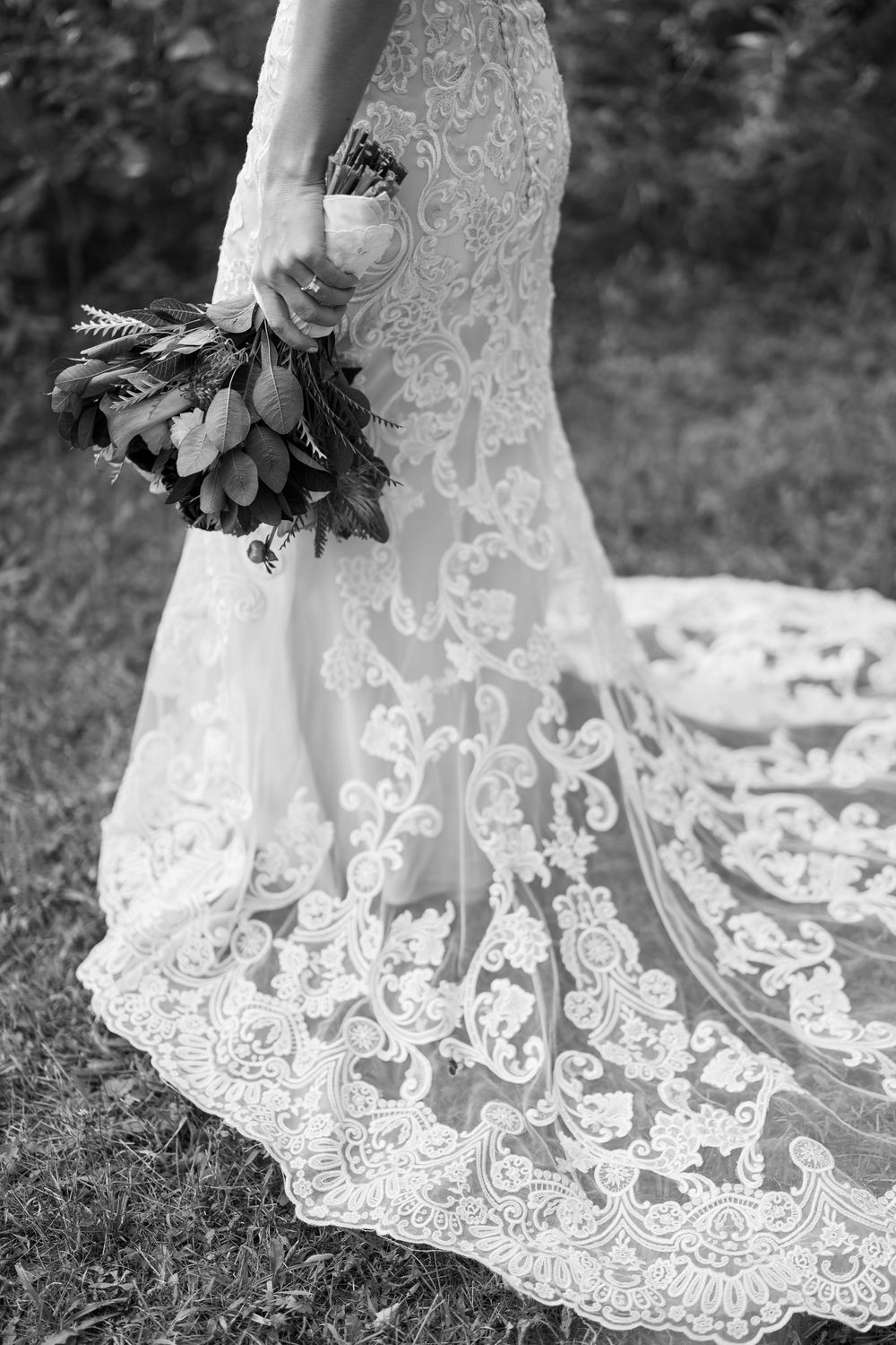 Black and white photo of a bride in a lace wedding gown with a long train, holding a floral bouquet outdoors.