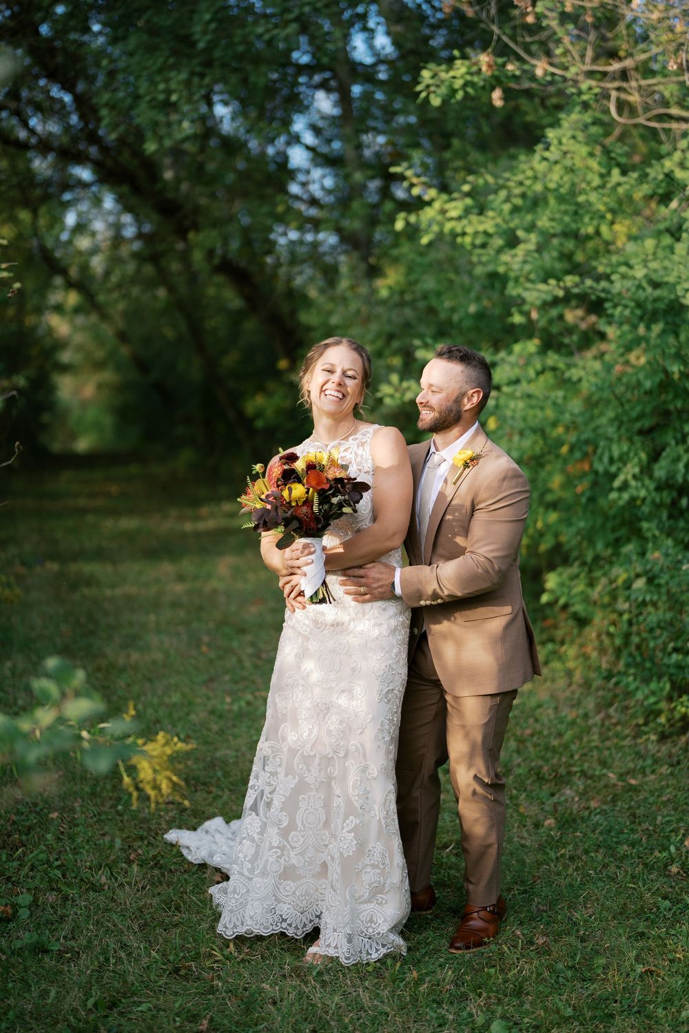 A bride and groom smile together outdoors in lush greenery, she in lace gown holding fall bouquet, he in tan suit.