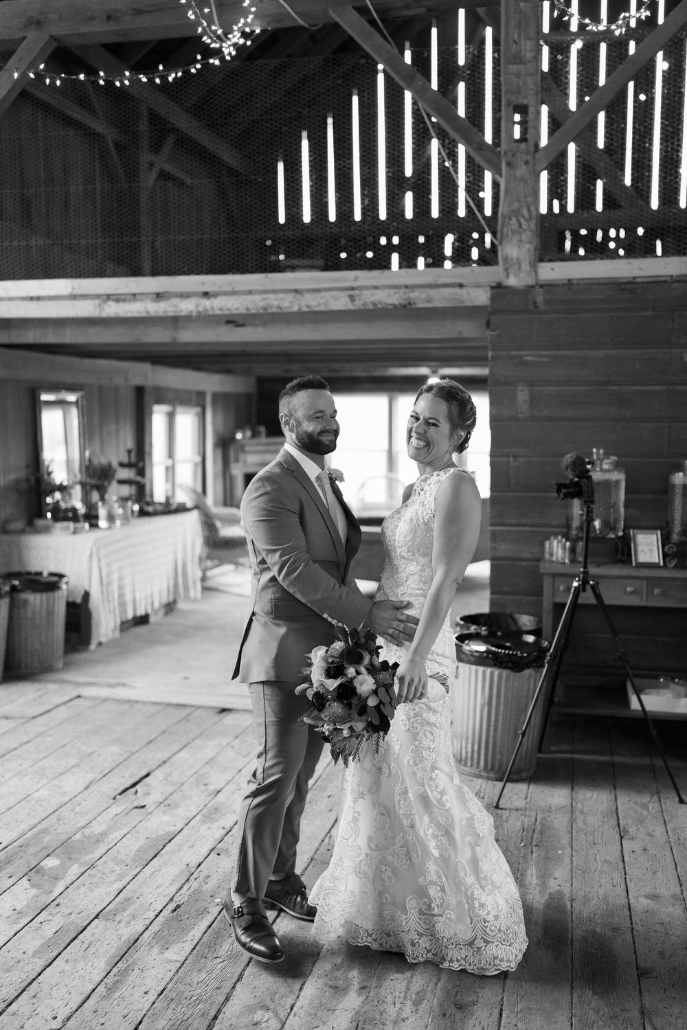 Black and white photo of a smiling bride and groom sharing a tender moment inside a rustic barn wedding venue.