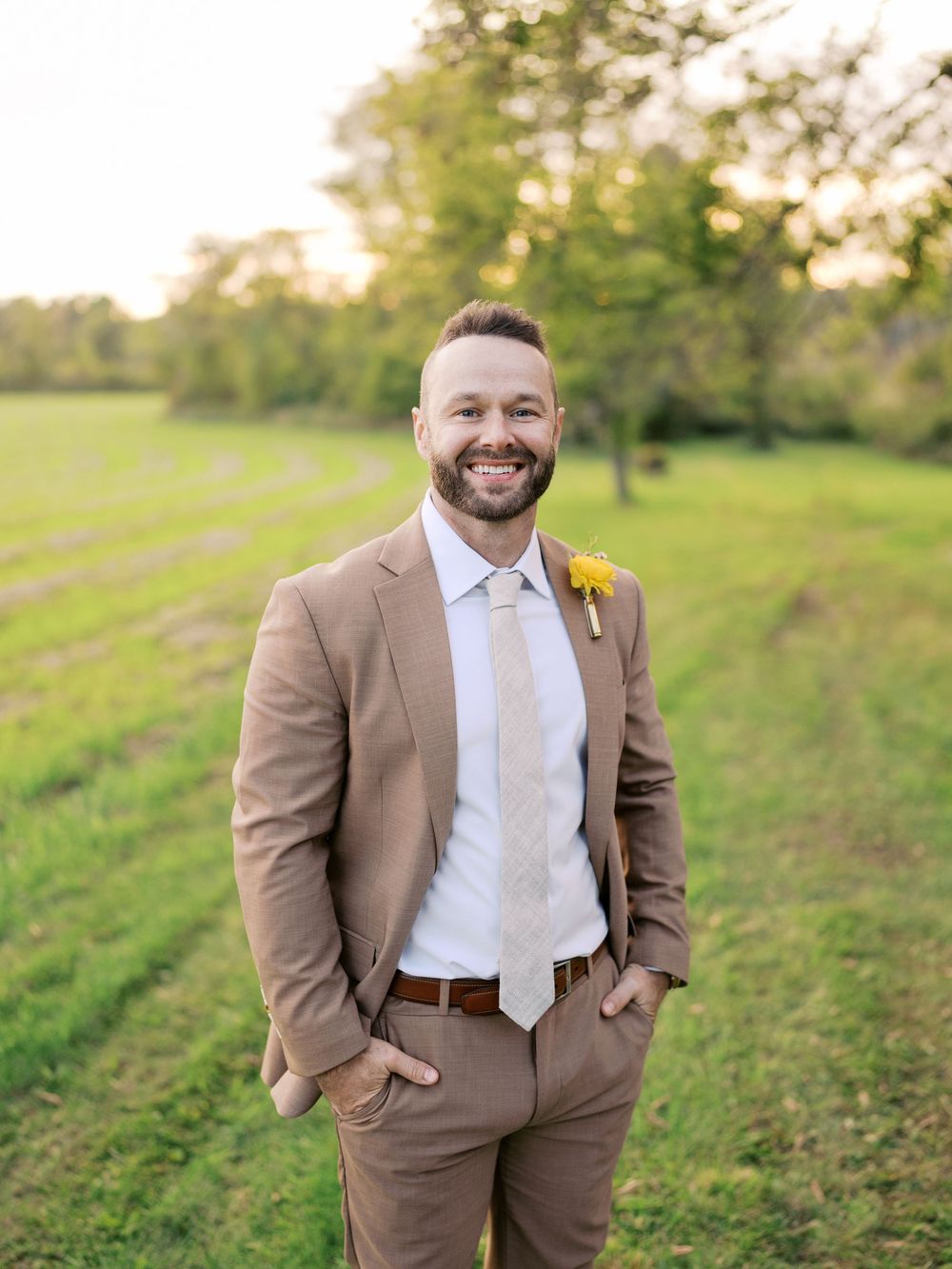 A smiling groom in a tan suit with yellow boutonniere stands in a lush green field at golden hour.