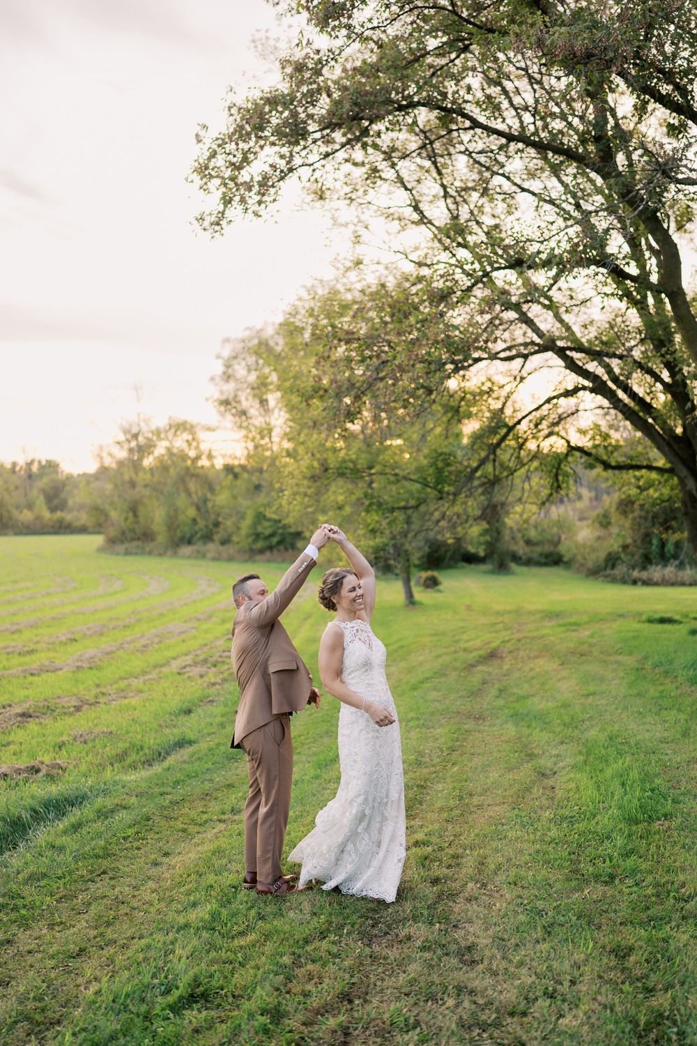 Bride and groom dancing outdoors at sunset in a lush green field, she in lace gown, he in tan suit.