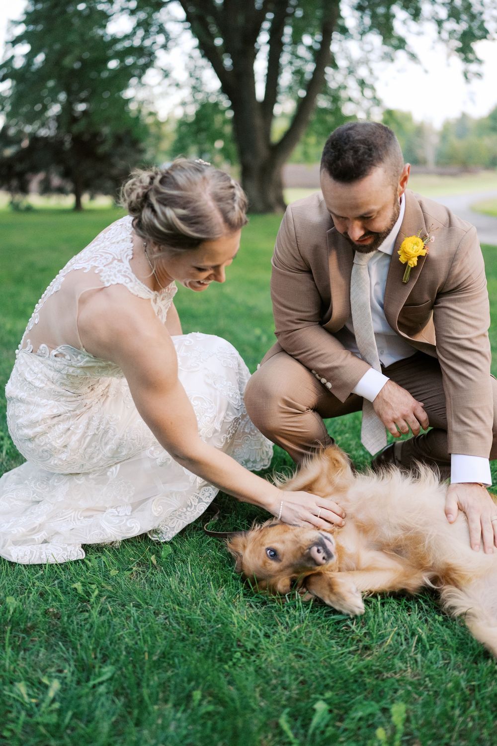 Bride and groom in wedding attire petting a golden retriever dog on green grass outdoors.