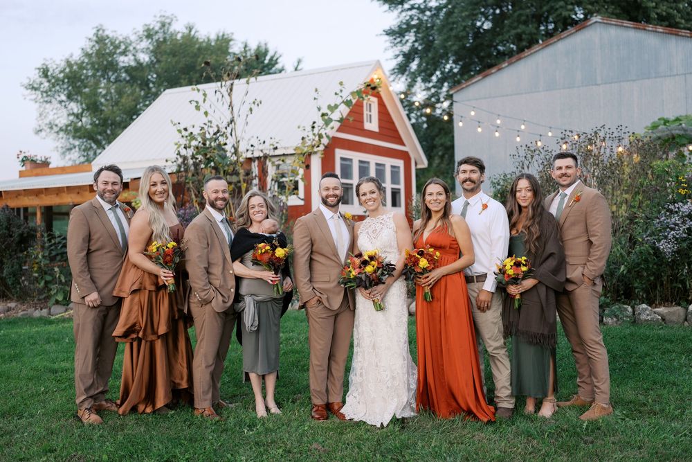 Wedding party poses in fall attire with bouquets outside a red barn venue on a sunny day.