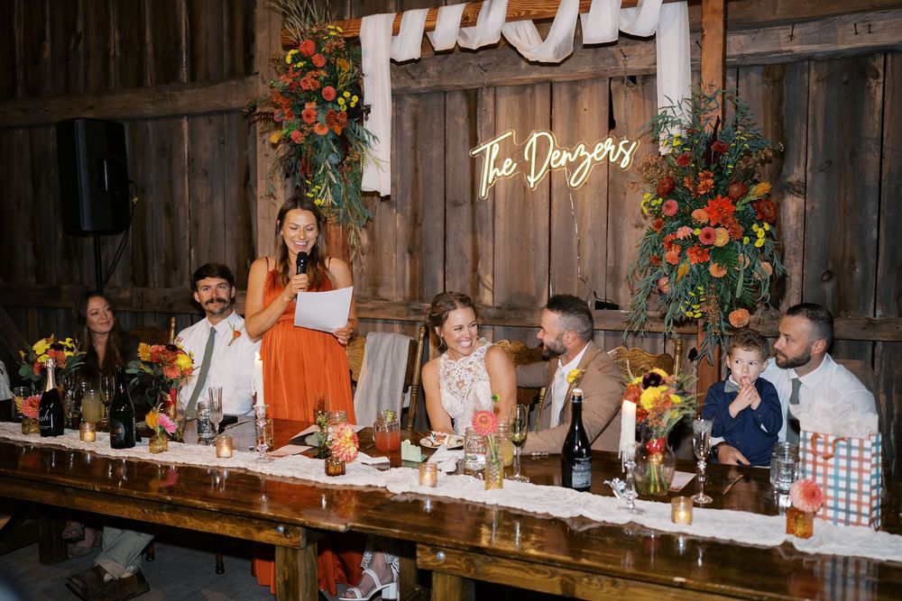 Wedding reception in a rustic barn with bridal party at a long decorated table, colorful floral arrangements, and neon sign.