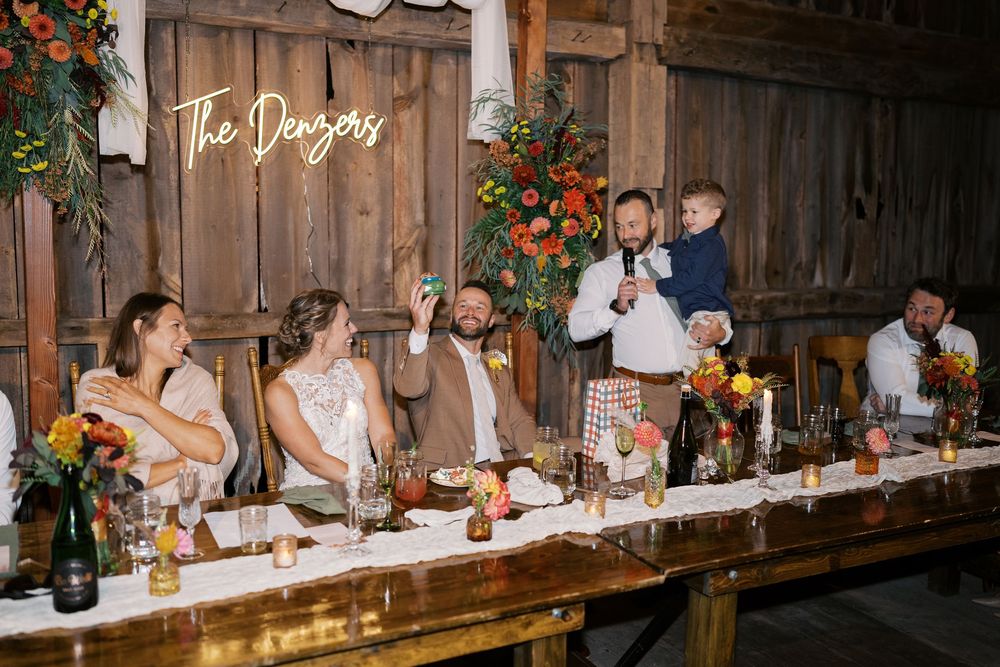 Wedding reception toast at rustic barn venue with floral arrangements, head table guests, and neon sign reading The Denzers.