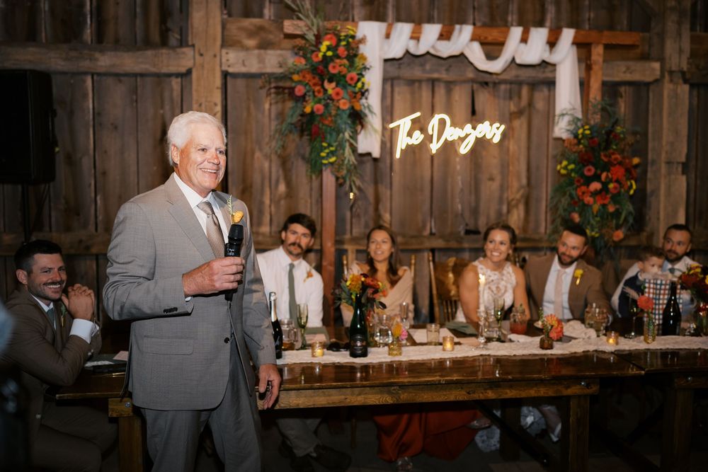 Father of the bride gives a heartfelt wedding toast in a rustic barn venue with floral decor.