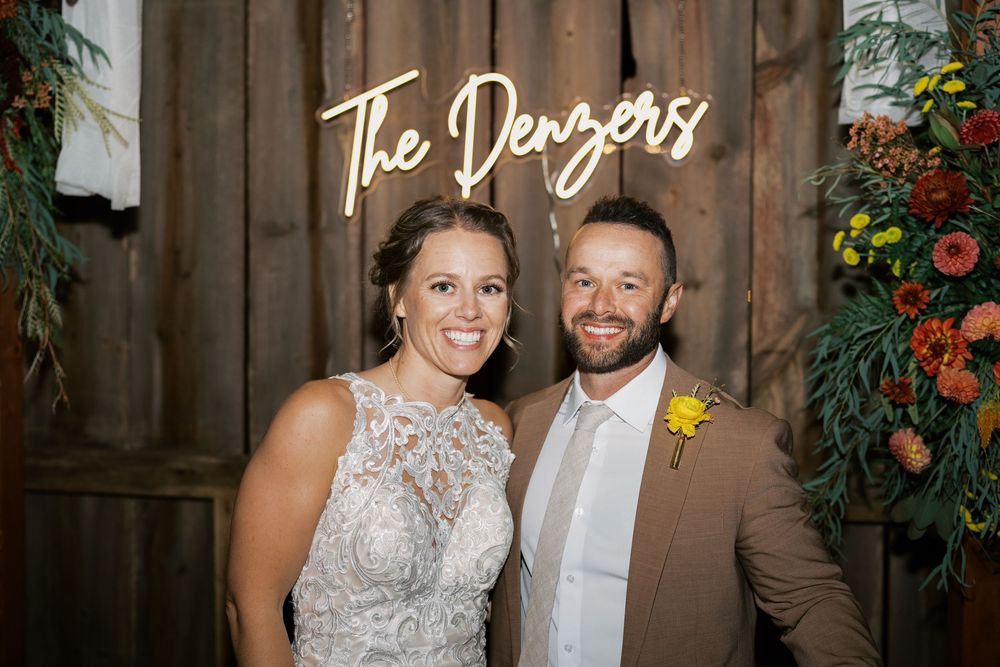 A smiling bride and groom pose under a neon sign reading The Denzers at their rustic barn wedding reception.