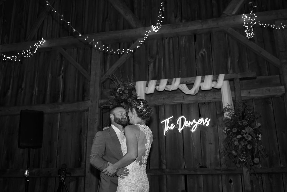 Black and white photo of newlyweds sharing first dance in rustic barn with neon sign reading The Danvers.