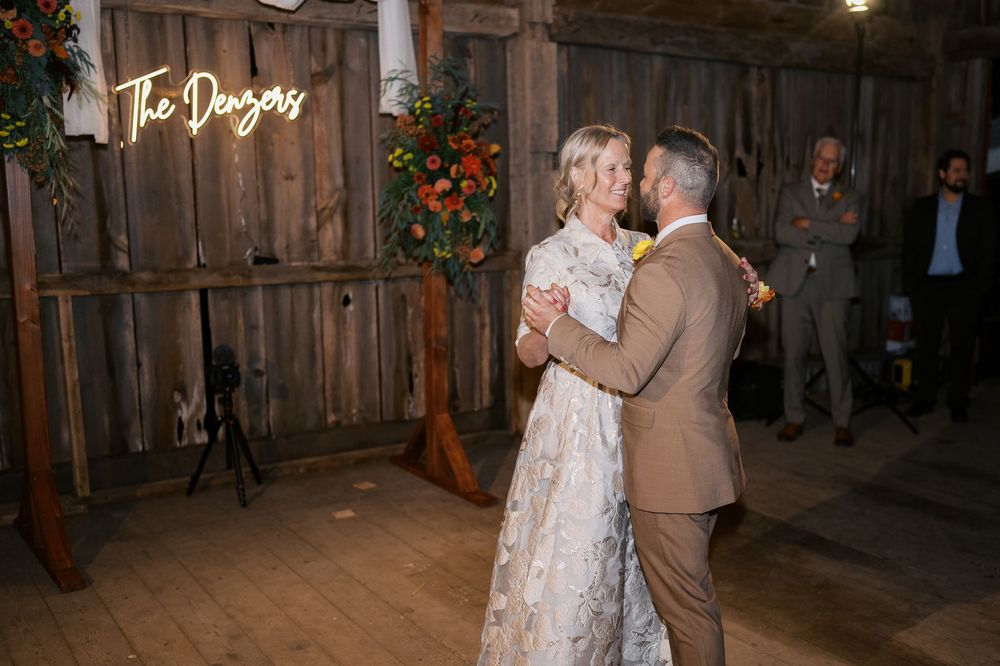 Newlyweds share their first dance in a rustic barn venue with a glowing neon sign reading The Dangers.