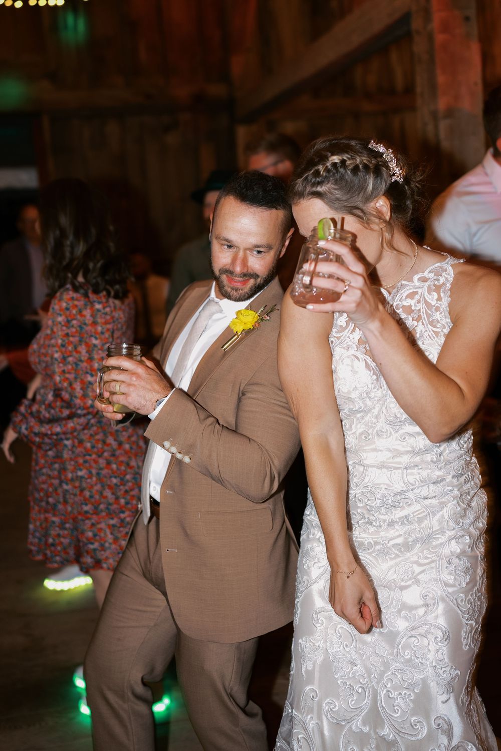 Bride and groom dancing together at wedding reception, holding drinks, groom in tan suit with yellow boutonniere.