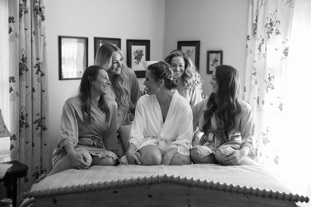 Five bridesmaids in robes laughing together on a bed in black and white wedding preparation photo.