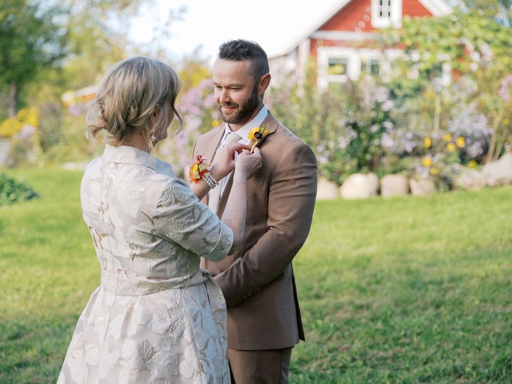 A woman pins a floral boutonniere on a smiling groom in a brown suit outdoors near a red barn.