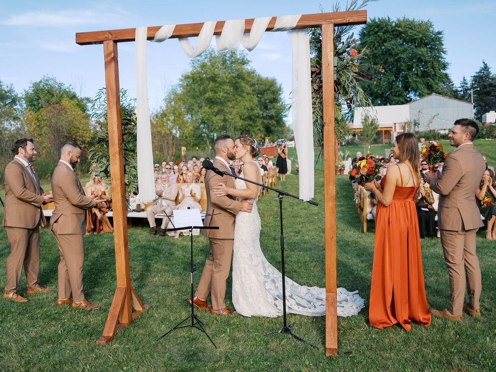 Couple kissing at outdoor wedding ceremony under wooden arch with wedding party in tan and orange attire on green lawn.