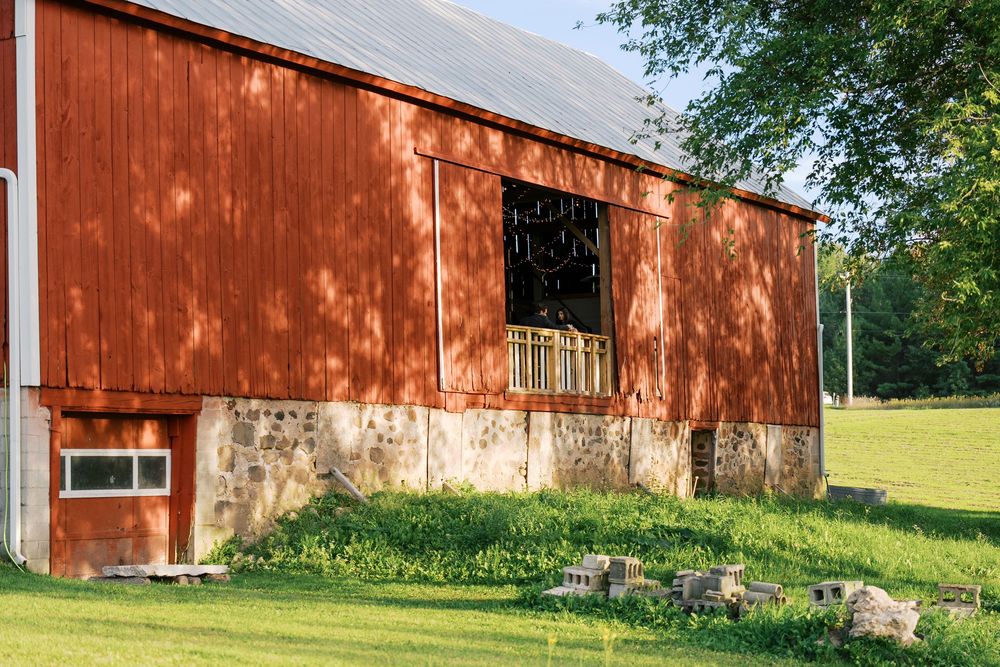 Red barn with stone foundation and open hayloft window surrounded by green grass and trees on a sunny day.