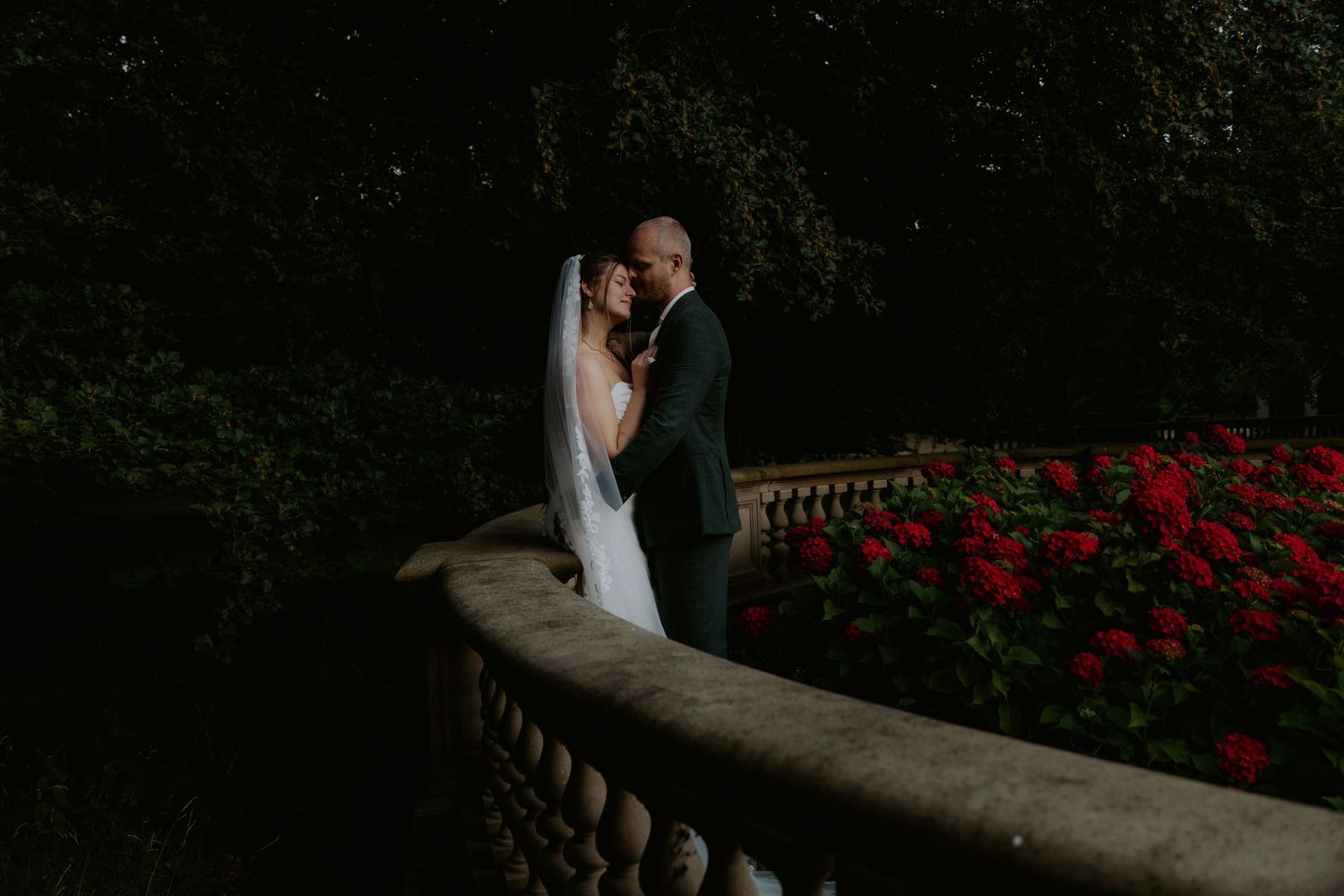 Bride in white lace wedding gown and cathedral veil stands before a sunlit window on herringbone hardwood floors.