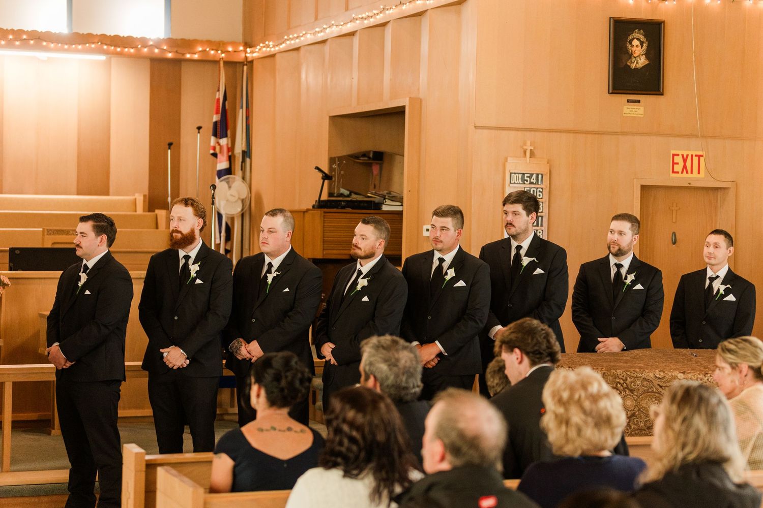 Groomsmen in black suits with boutonnieres stand in a row at a church wedding ceremony.