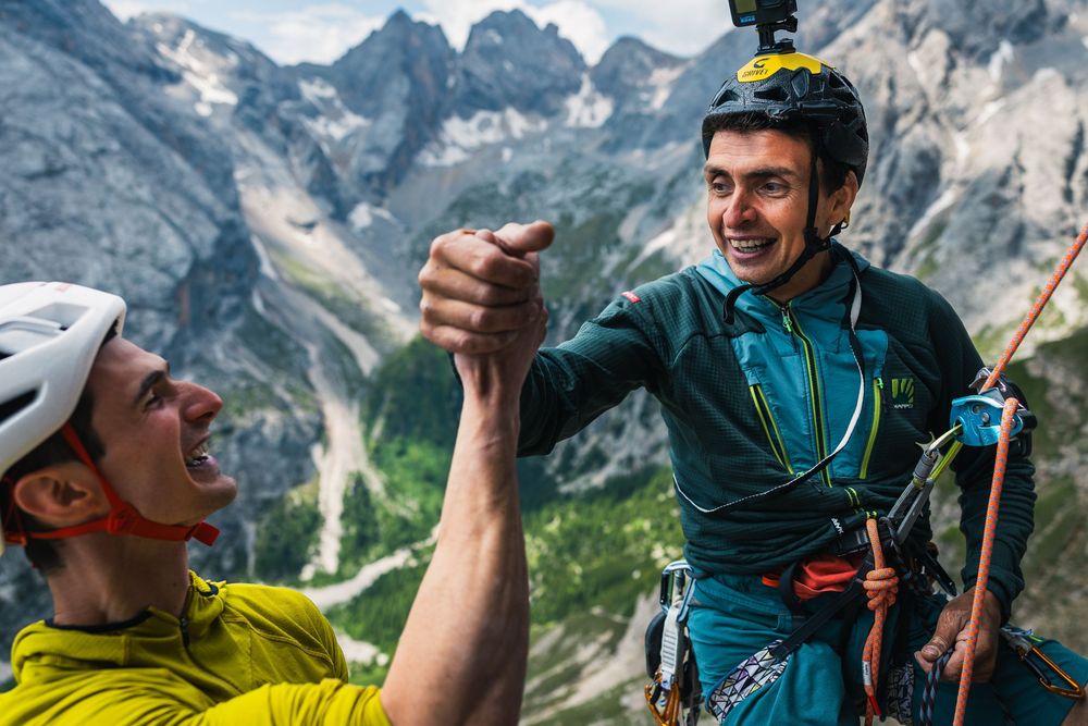 Adam Ondra and Berni Rivadossi shake hands at a belay on Egoland, with Dolomite peaks behind. Photo: Jan Virt
