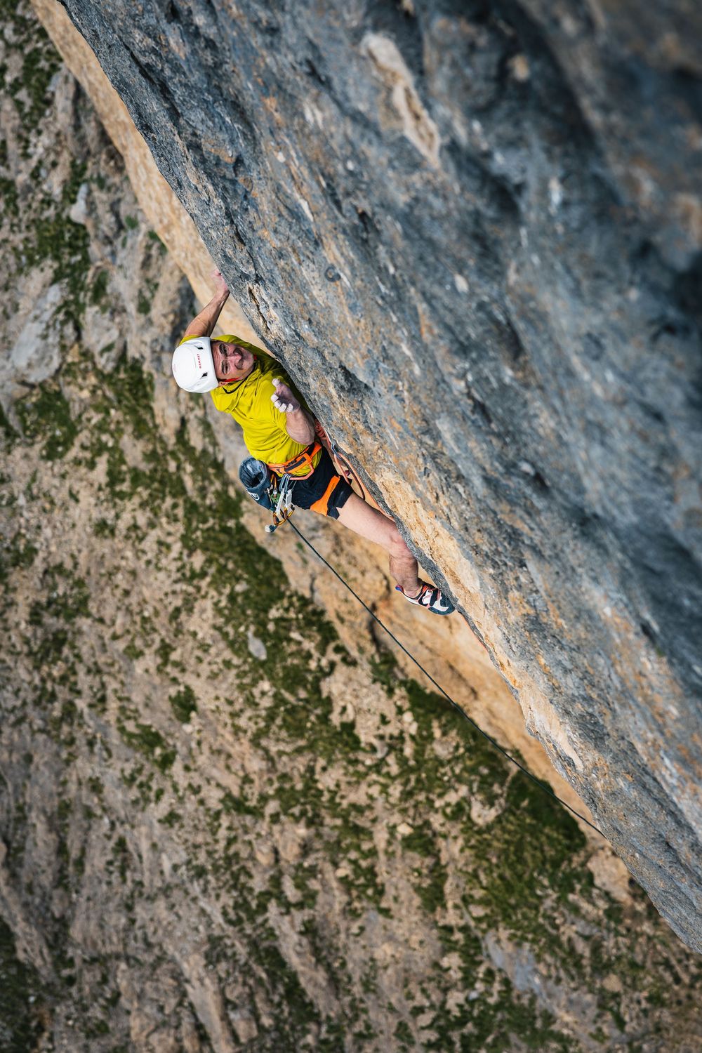 Adam Ondra in a yellow top and white helmet on a steep grey slab of Egoland above a scree basin, Marmolada. Photo: Jan Virt
