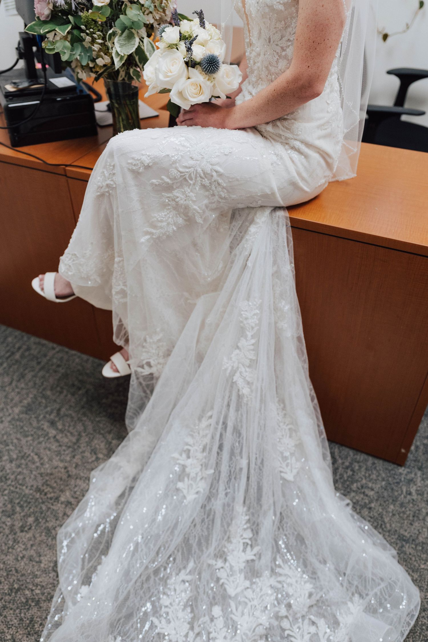 Bride in lace wedding gown with floral train sits on desk holding white bouquet with blue thistle accents.