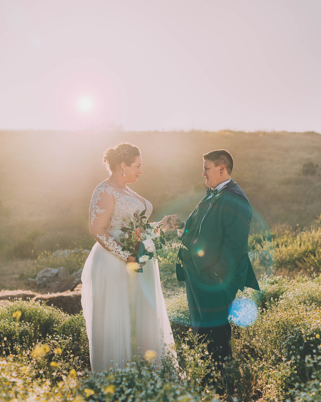 Bride and groom exchange vows outdoors at golden hour, surrounded by wildflowers with sunlight glowing behind them.