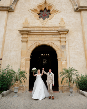 Bride and groom celebrating at ornate church entrance, bride in white gown holding bouquet, groom in tan suit.