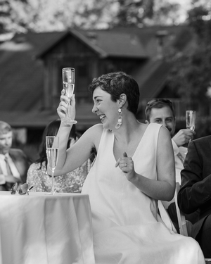 Black and white photo of a bride raising a champagne glass and laughing during a wedding toast outdoors.