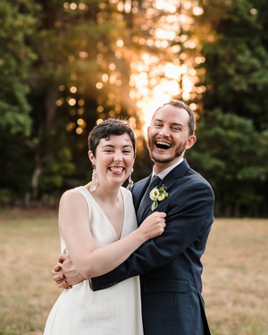 A joyful couple in wedding attire laughing together outdoors at golden hour with trees in the background.
