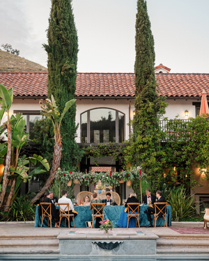 Elegant outdoor wedding reception at a Spanish-style villa with blue velvet tables, tall cypress trees, and pool.