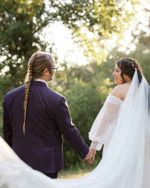 A bride and groom hold hands outdoors, she wears a veil and off-shoulder gown, he wears a navy suit with a long braid.