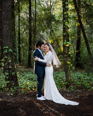 Bride and groom embrace in a lush forest setting, wearing white lace gown with veil and navy suit.