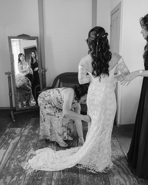 Black and white photo of a bride in lace gown being helped by bridesmaids before wedding ceremony.