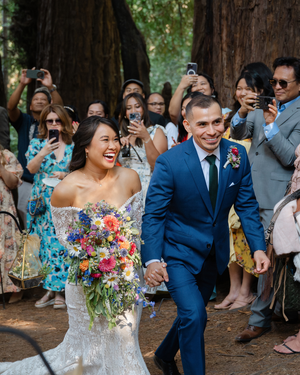 Joyful bride and groom walk through redwood forest ceremony aisle, guests cheering, bride holding colorful wildflower bouquet.