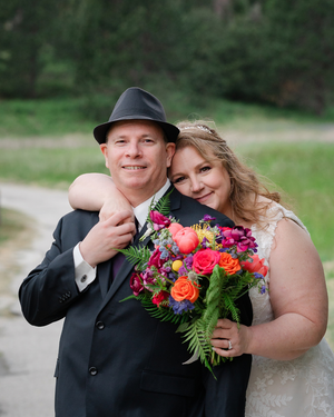 Bride and groom pose together outdoors, she holds a vibrant bouquet of pink, orange, and purple flowers, he wears a black hat.